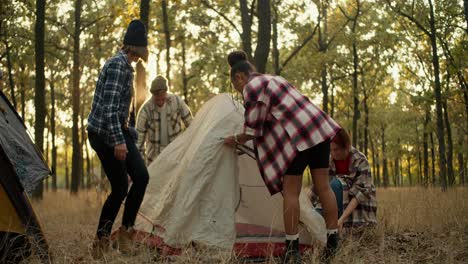 A-girl-with-black-skin-in-a-checkered-shirt,-along-with-the-rest-of-the-expedition-members,-sets-up-her-white-tent-for-an-overnight-stay-in-a-green-summer-forest