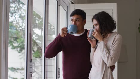 Romantic-hispanic-couple-embracing-standing-in-window-having-coffee