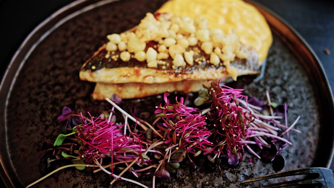 Primo piano di un filetto di pesce bianco scottato in padella con pelle croccante, guarnito con cereali soffiati croccanti.