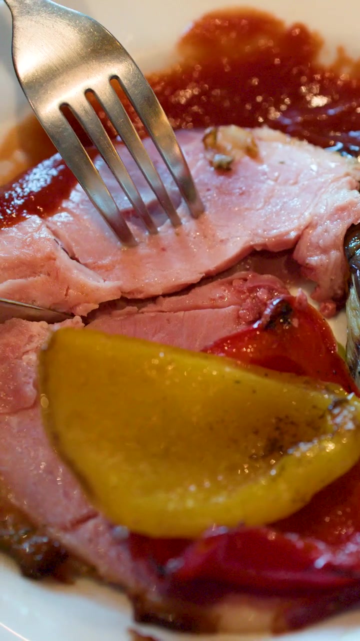 Close-up shot of a fork and knife cutting roasted ham served with grilled bell peppers