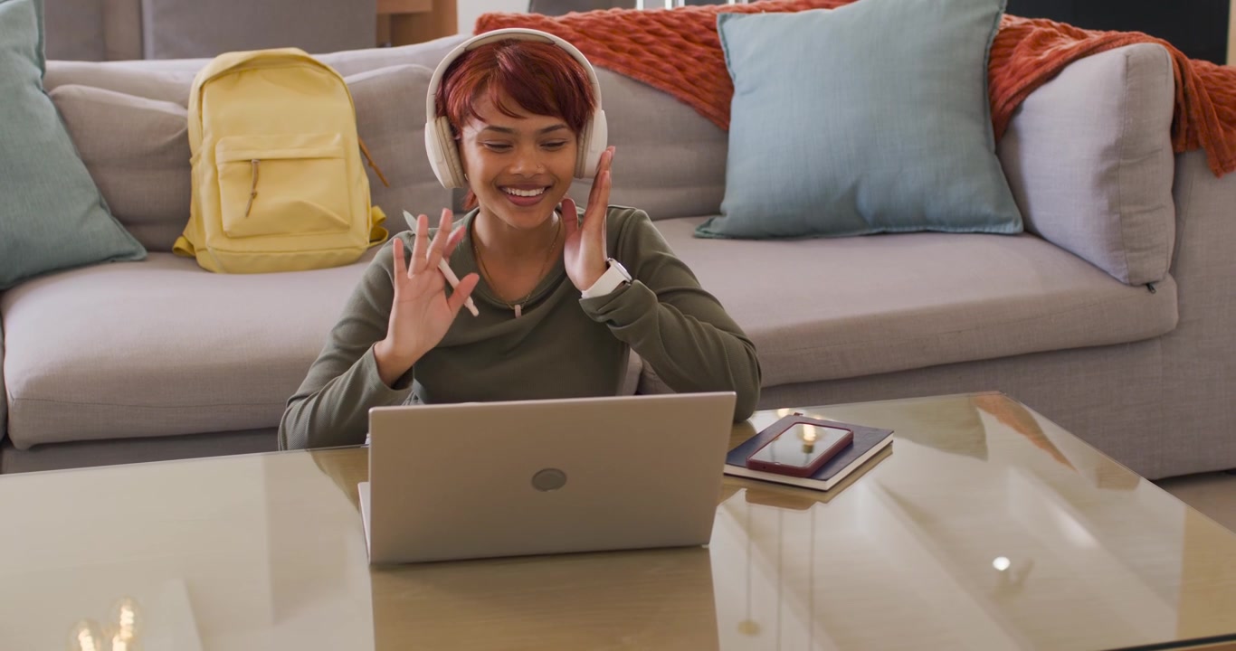 African American woman receiving invite adjusting headset waving at laptop noting stylus at home