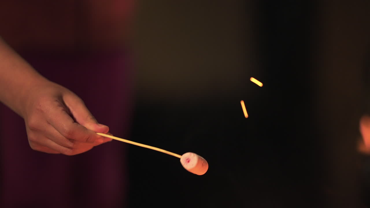 Static close-up captures a marshmallow toasted over a crackling fire, held by hand gripping a wooden skewer, begins to brown as bright orange flames flicker nearby