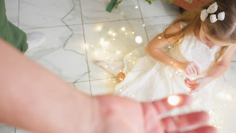 hand view holding and arranging christmas lights, while daughter assists by sitting on floor, helping with decoration around festive tree, creating cozy holiday atmosphere