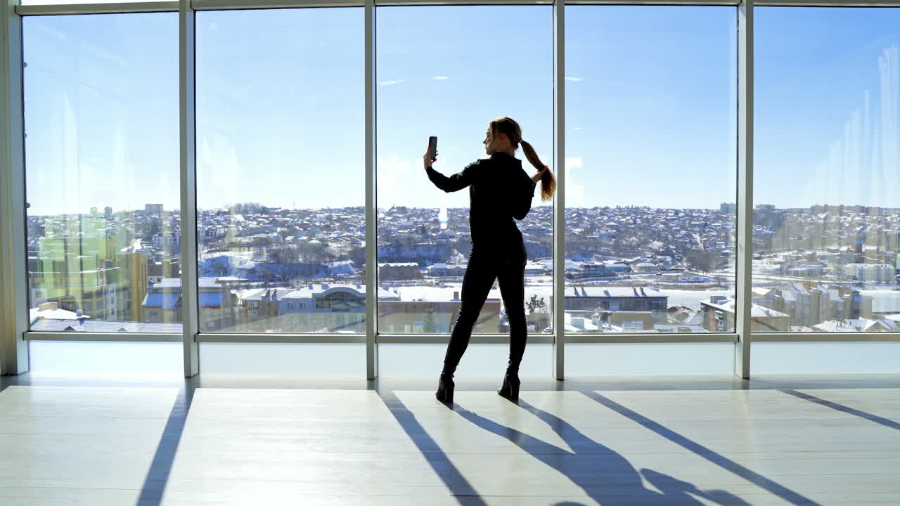 Young woman taking selfie photo in the office. Beautiful girl standing by the large panoramic windows and taking photo of herself on a phone. Rear view.