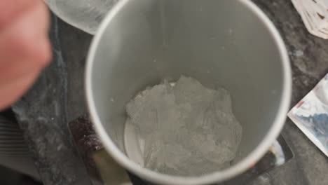 close up of translucent rubber like soluble material inside metal jug with person dropping more pieces into jug while working on project on marble counter, indoor setting with scattered supplies