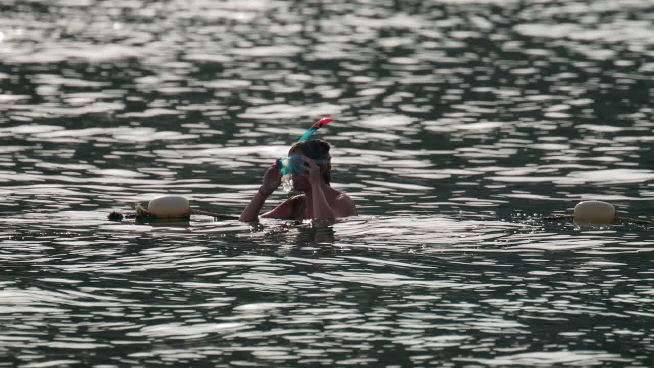 A woman prepares for snorkeling in the ocean, adjusting her mask while floating in gentle water under soft sunlight