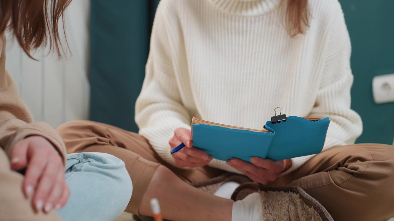 Sessione di tutoraggio a domicilio con quaderno blu, due persone sedute a gambe incrociate sul pavimento in una stanza accogliente, il tutor che guida lo studente attraverso esercizi di grammatica, una penna che batte sulle pagine, maglioni casual, luce naturale calda.