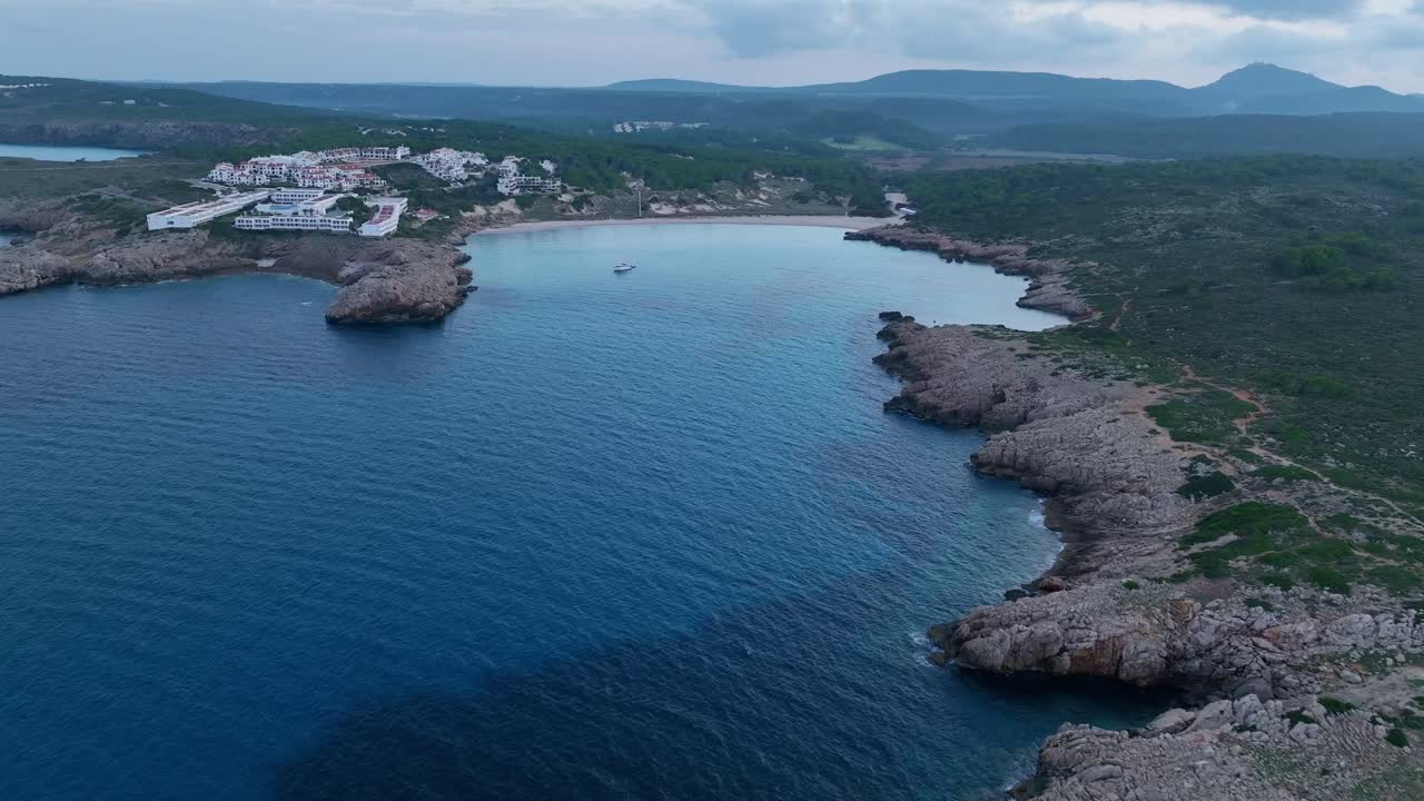 Filmisk droneflyvning over en lille by på Menorca, Spanien, med en strand med krystalklart blåt vand.