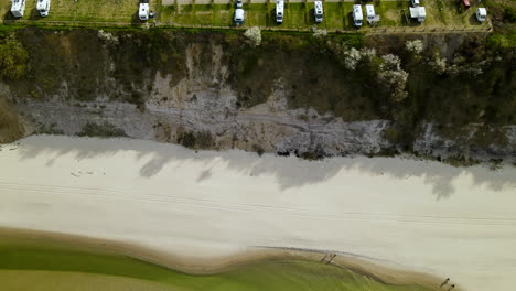 Aerial---Drone-flying-away-from-a-baltic-sea-white-sand-beach-showing-the-parking-lot-for-camper-trailers-on-the-cliff-in-Chlapowo,-Poland