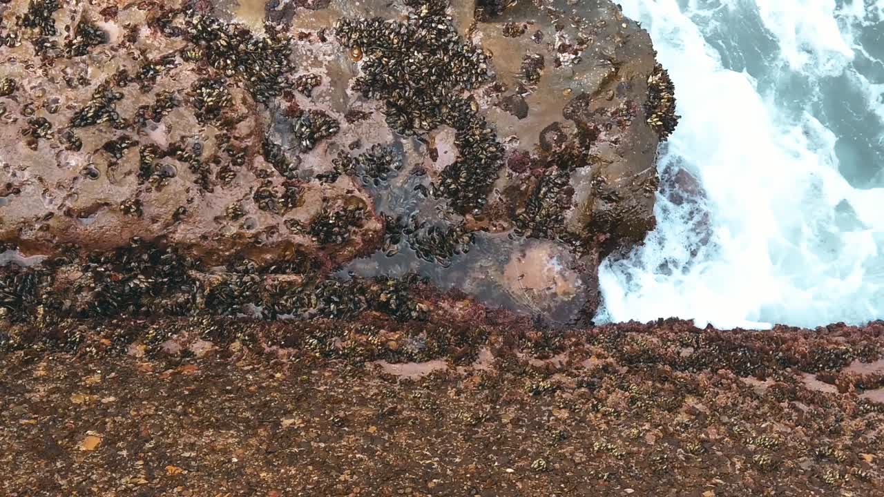 Blue ocean waves flowing on a big brown rock with little stones. Overhead view in daylight. Slow motion