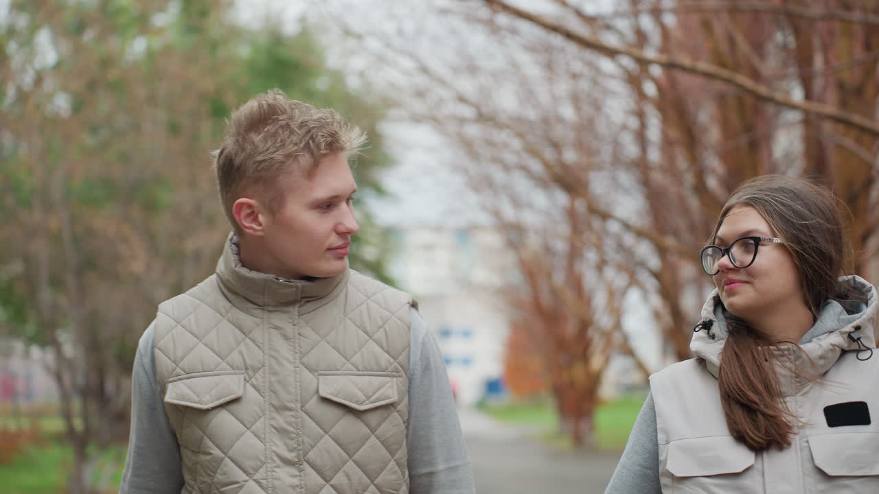 Close moment of friends in matching casual outfits warmly staring at each other outdoors with soft smiles, surrounded by greenery and buildings