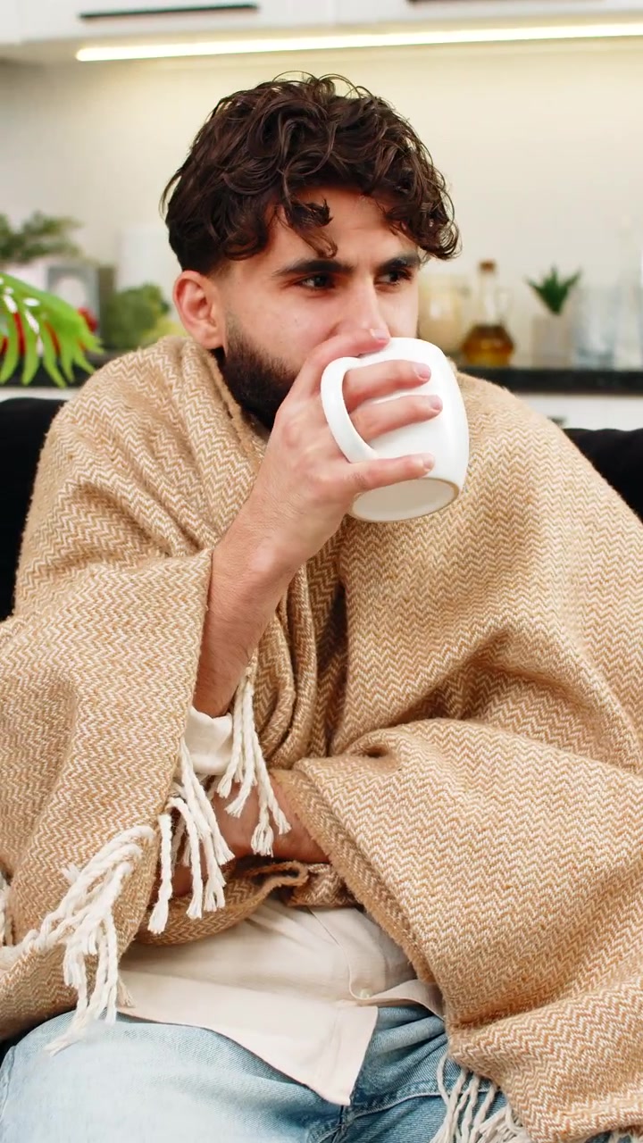 Uomo indiano avvolto in una coperta, che tiene in mano una tazza di tè caldo, bevendo lentamente e rilassandosi dopo il lavoro, sorridendo.