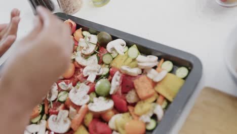 Hand-of-biracial-woman-seasoning-chopped-vegetables-on-baking-tray-in-kitchen,-slow-motion