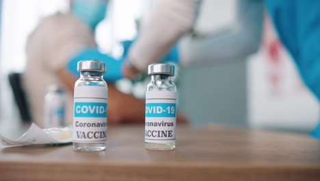 close up shot of coronavirus vaccine ampoules with liquid, ampoule with vaccine from covid 19 on table in clinic room, doctor making injection on background, injection set, vaccination concept