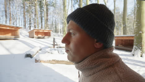 primer plano de un hombre en traje de invierno bebiendo de una botella térmica en un bosque nevado con cabañas de madera en el fondo
