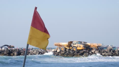 red, yellow flag waving in the wind on a stormy beach in front of a yellow truck on a construction site in dubai