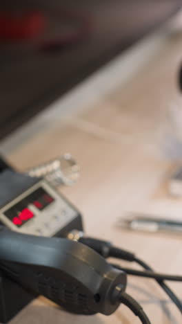 una vista en primer plano de una mano con un guante negro que opera una estación de soldadura en un laboratorio, la configuración incluye un microscopio con un circuito debajo y varias herramientas electrónicas en un escritorio de madera