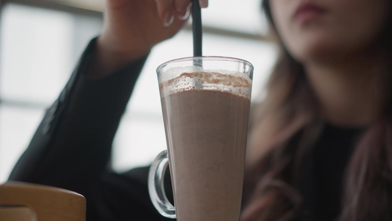 Primo piano di una giovane donna che tiene una tazza alta e trasparente di milkshake al cioccolato su un tavolo di legno, mescolando con una cannuccia nera e portando la tazza alla bocca in una morbida luce naturale da caffè.