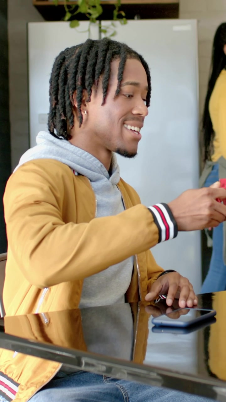 Vertical video: Smiling man in yellow jacket lifting red mug at glass table as woman passes, phone