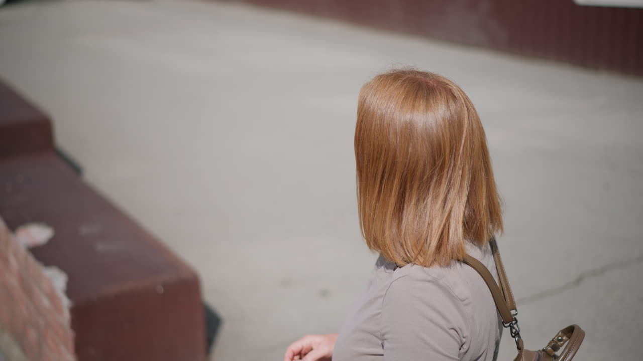 Aerial view of solitary muse standing under strong summer sunlight near building, looking around for someone and checking time with tired expression, sunlight on face