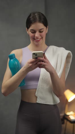 vertical video confident and happy sporty brunette girl in a purple top writes on social networks using a white smartphone holds a white towel and a blue sports bottle in her hands after playing sports in a modern apartment