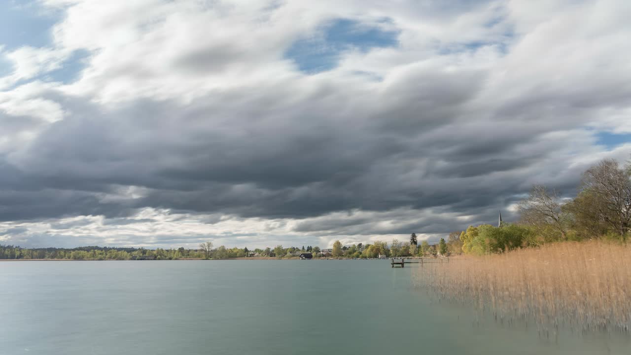 Dark clouds drift over a lake. Nature is beautiful, and the landscape is blooming, for it is spring. The sun illuminates the plants and the lake.
