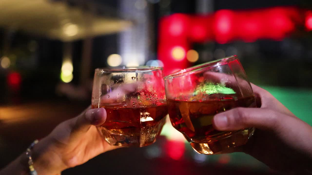 Two people clink cocktail glasses in a celebratory toast beside a brightly lit rooftop pool in Bangkok, with colorful evening lighting and shallow depth of field