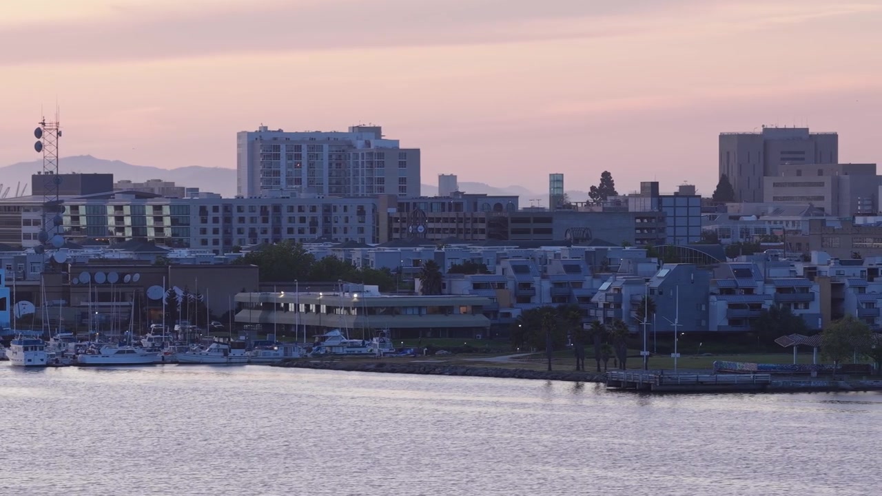 Cielo rosa al tramonto sul lungomare di Oakland, con una splendida vista sullo skyline della città, la marina di Jack London Square e moderni edifici che riflettono l'ultima luce lungo le calme acque dell'estuario.