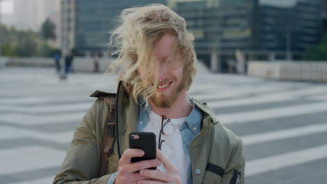 retrato de un joven atractivo turista que usa un teléfono inteligente disfrutando de mensajes de texto navegando por mensajes en línea compartiendo experiencia de viaje sonriendo feliz en la ciudad el viento soplando el cabello en cámara lenta