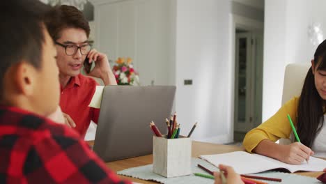 Asiatischer-Vater-Nutzt-Smartphone-Und-Laptop-Und-Arbeitet-In-Der-Küche,-Während-Sohn-Und-Tochter-Schularbeiten-Erledigen