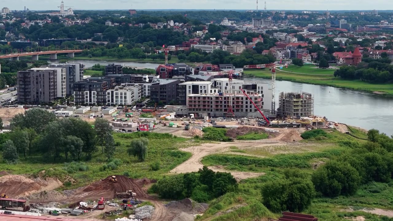 Luchtopnamen tonen een bouwplaats aan de rivier met kranen, nieuwe appartementencomplexen en weelderig groen, tegen een levendige stad onder bewolkte daglichtlucht