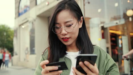 close up of the young beautiful girl in glasses standing at the street with a credit card in hand and shopping online on the smartphone