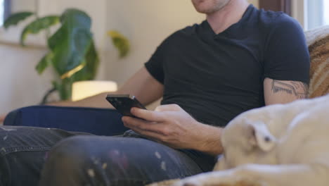 a man sits on his couch with his dog and scrolls on his phone wasting time