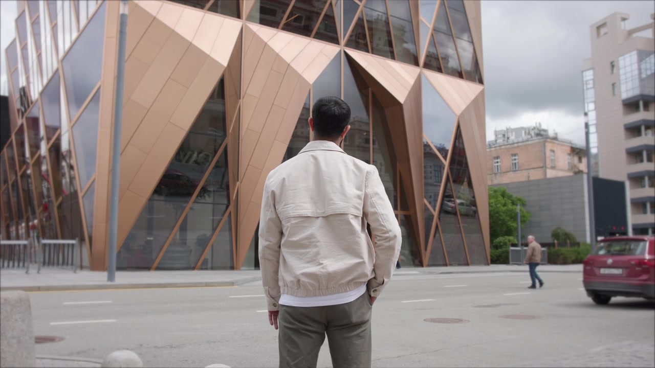 Hombre visto de espaldas de pie en la acera frente a un gran edificio comercial contemporáneo con paneles metálicos de bronce angulares y altas ventanas de cristal reflectantes mientras el tráfico circula junto a un peatón cercano.