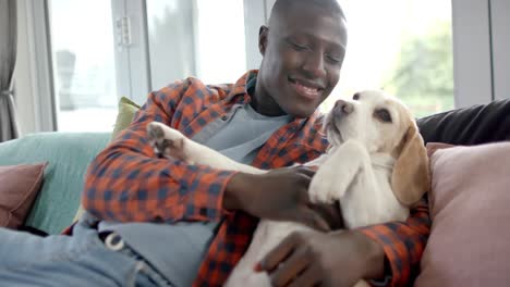 happy african american man sitting on sofa, embracing his pet dog at home, slow motion