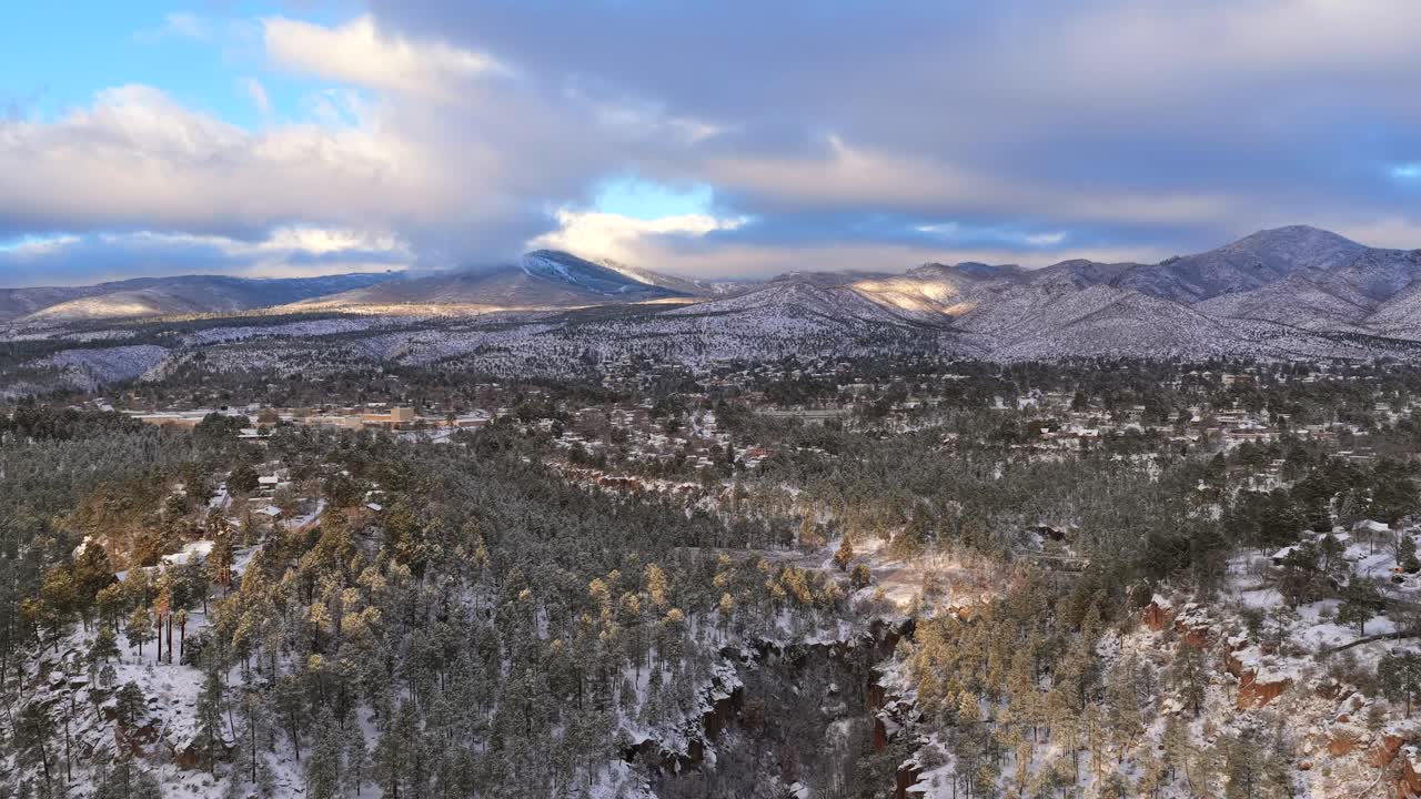 Expansive view of a town built along the edge of a deep, snow-dusted canyon. Captures the dramatic winter sunrise aesthetic in the high-altitude Southwestern pine forest