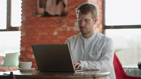 adult man in white shirt and eyeglasses is sending message in online chat by modern laptop working in cafe
