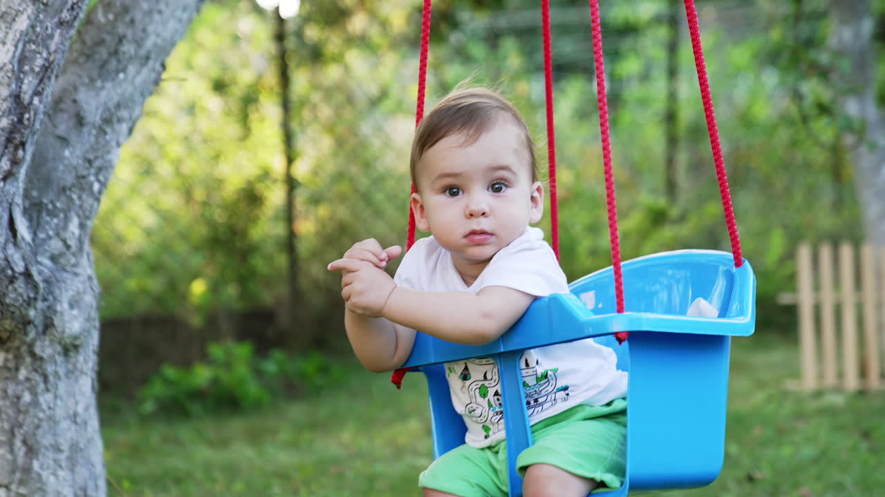 Sweet Caucasian baby boy with bare feet sits in a swing. Cute child having good time outdoors in summer. Blurred nature backdrop.