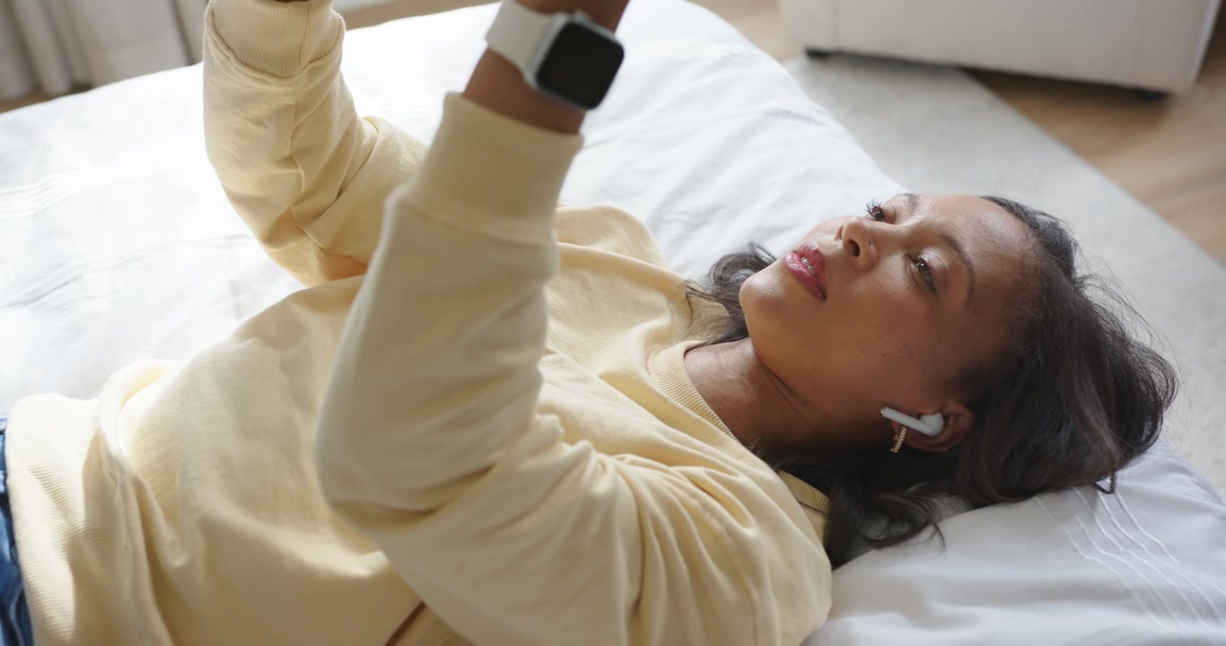 Donna africana adulta sdraiata sul letto, inserisce un auricolare bianco e solleva un dispositivo nero sopra di sé per guardarlo.