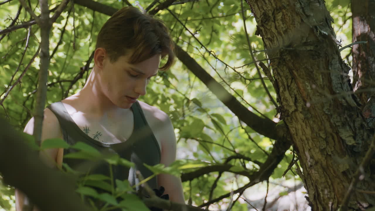 tourist under dense foliage reaches into bag by large barked tree trunk with focused expression amid sunlit green canopy and forest silence suggesting resource search and cautious exploration
