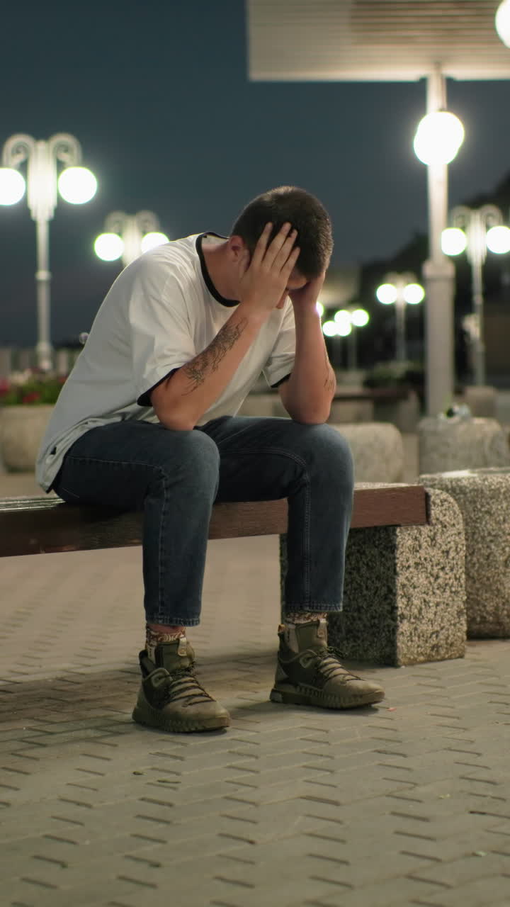 hombre sentado en un banco al aire libre por la noche con las manos cubriendo su cara y quitándola, pareciendo estresado o ansioso, las luces de la ciudad y los peatones lejanos crean una atmósfera urbana tranquila en el fondo