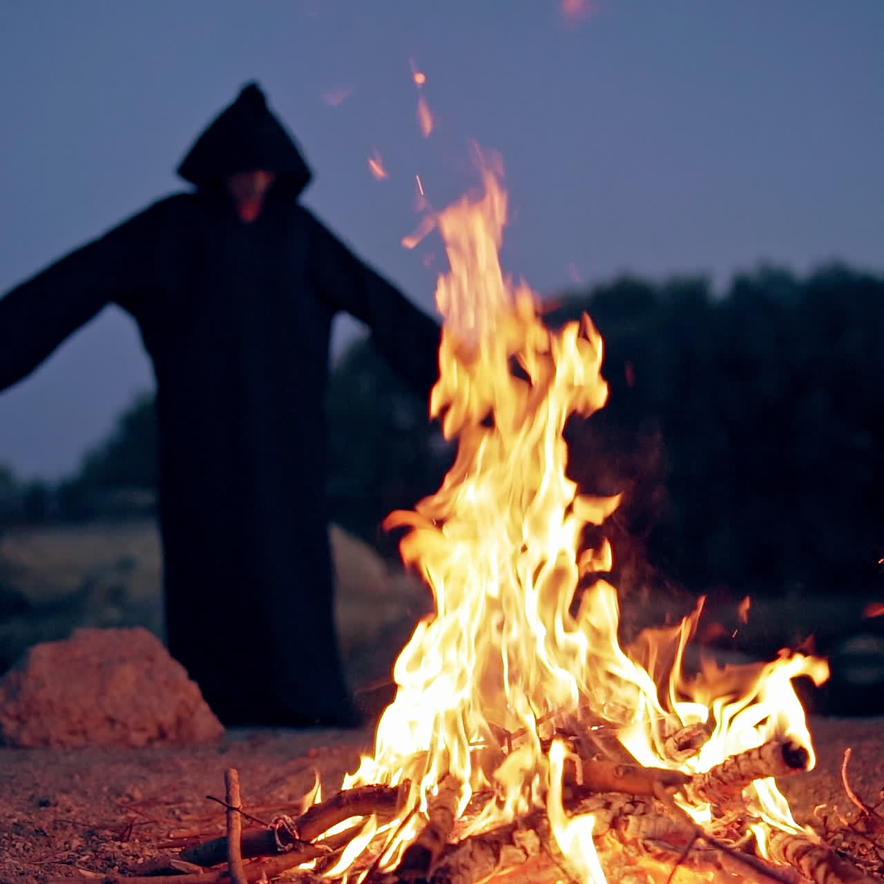 Silhouette of black death by the fire at night with hands outstretched. Freedom concept. Flame on the ground and witch costume on the natural background in the evening.