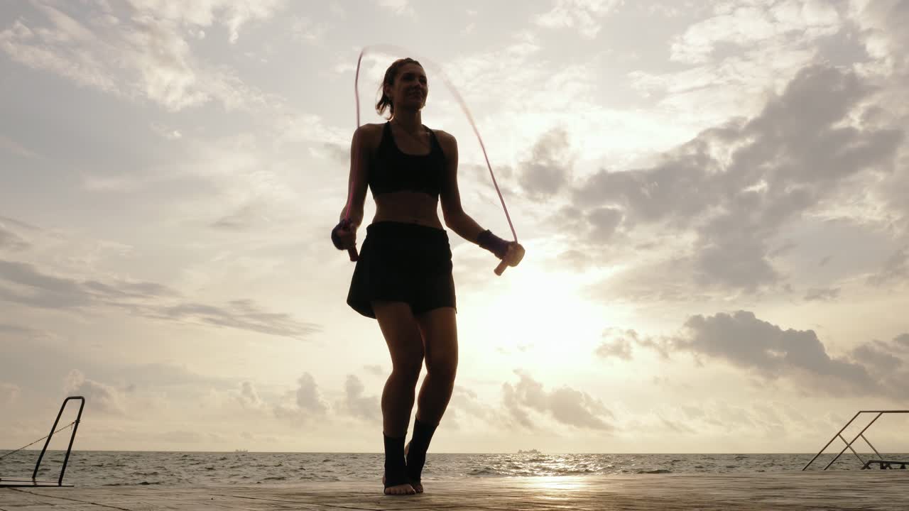 View form the bottom: Young woman working out on the jump rope against the sun by the beach in slowmotion. Lens flare. Girl jumping on a skipping rope by the sea. Shot in 4k