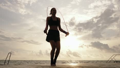 Ansicht-Von-Unten:-Junge-Frau-Trainiert-Am-Strand-In-Zeitlupe-Am-Springseil-Gegen-Die-Sonne.-Linseneffekt.-Mädchen-Springt-Auf-Einem-Springseil-Am-Meer.-In-4k-Gedreht