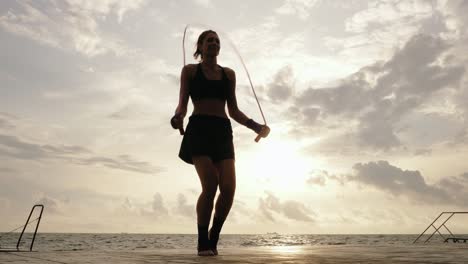 vista desde la parte inferior: mujer joven trabajando en la cuerda de salto contra el sol junto a la playa en cámara lenta. destello de la lente. niña saltando en una cuerda de salto junto al mar. filmado en 4k