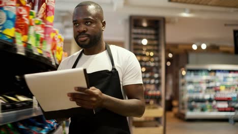 Un-Hombre-De-Piel-Negra-Con-Una-Camiseta-Blanca-Y-Un-Delantal-Negro-Camina-Por-Las-Ventanas-De-Un-Supermercado-Y-Hace-Inventario-Usando-Papel-Y-Una-Tableta