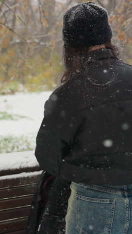 vista trasera de la dama abriendo el paquete de guitarra colocado en una silla cubierta de nieve en un parque de invierno sereno, con la nieve cayendo suavemente, una mezcla de árboles verdes y helados en el fondo