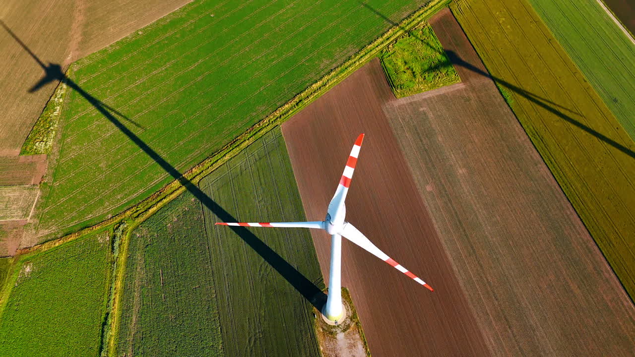 Wind turbine casting a shadow on fields. A wind turbine rises above lush green fields while casting a long shadow on the crops below during a sunny day