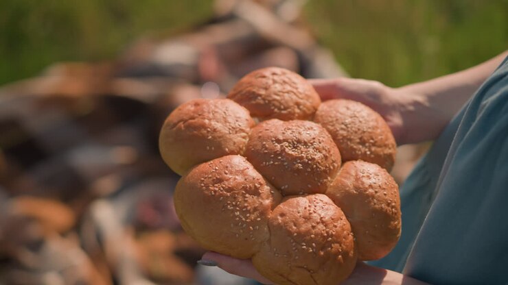 A close-up shot of a woman's hand in a blue dress holding a cluster of freshly baked sesame seed bread rolls. The background features blurred children. focused on the bread