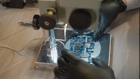 A close-up view of a technician in black hand gloves meticulously working on a circuit under a microscope in a laboratory